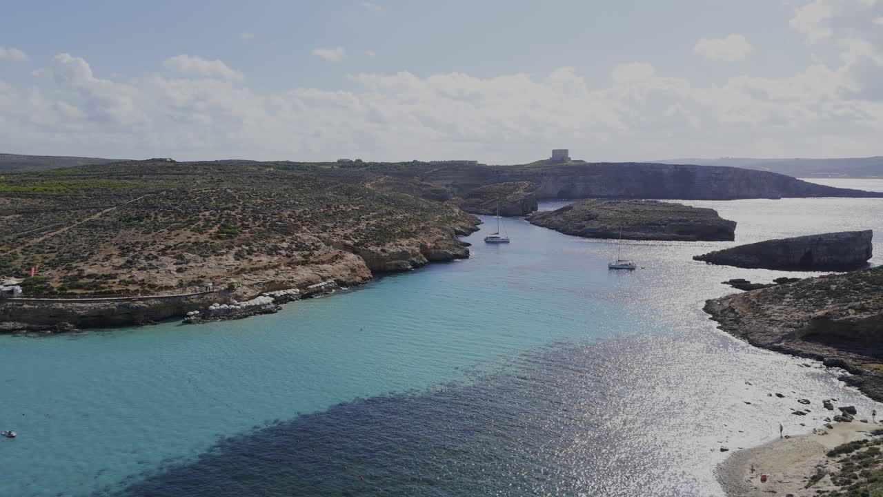 A calm panoramic drone shot over the Blue Lagoon shows Comino, Cominotto Beach and two yachts, with bright sunlight reflecting on clear turquoise Mediterranean water