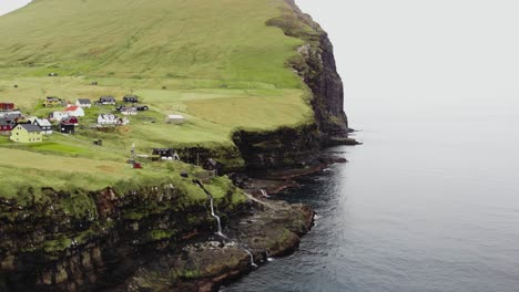 Antenne-Eines-Kleinen-Dorfes-Entlang-Der-Klippe-Eines-Kleinen-Berges-Mit-Wasserfällen-Und-Flüssen-In-Der-üppigen,-Mit-Gras-Bedeckten-Landschaft-Neben-Dem-Ruhigen-Nordatlantik