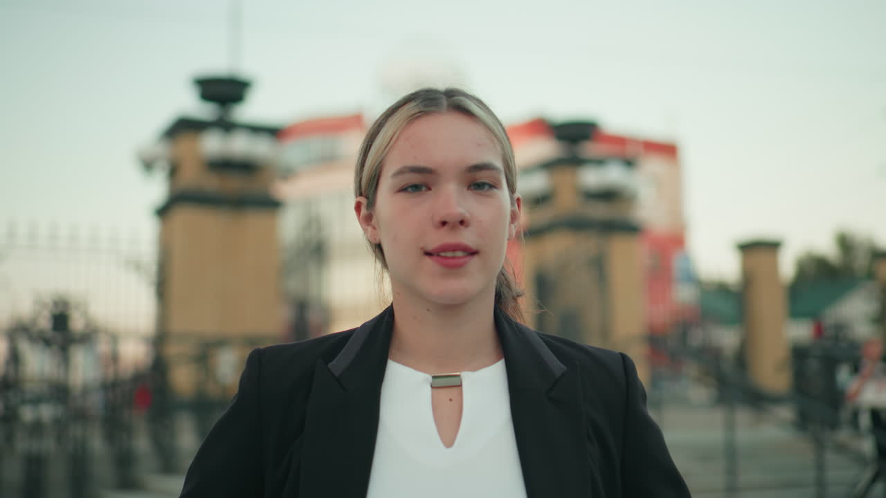 Elegant lady in professional attire stands confidently outdoors with hands on waist, exuding self-assurance in modern city setting with architectural background and people walking in blur behind her