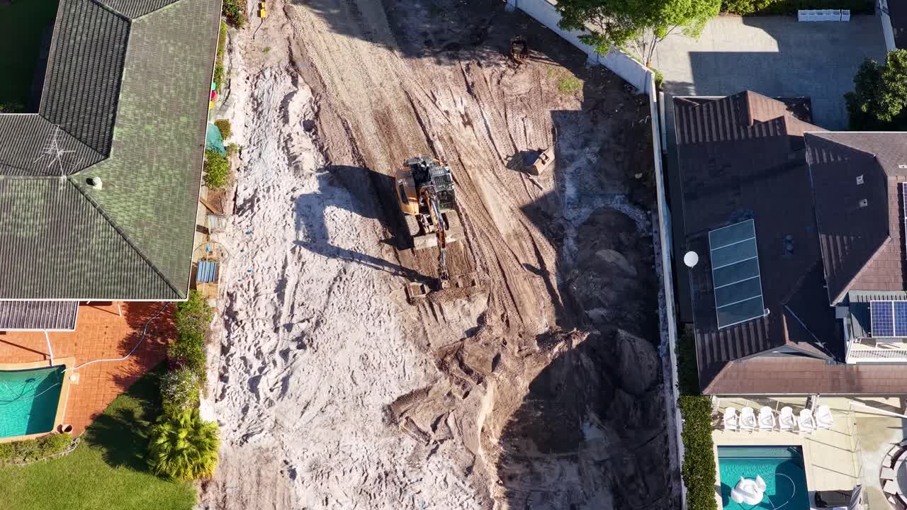 Excavator moves and loads dirt at suburban construction site, captured by drone in daylight