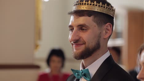 Groom-in-the-crown-stands-in-church.-He-looks-at-bride-and-smiles.-Newlyweds