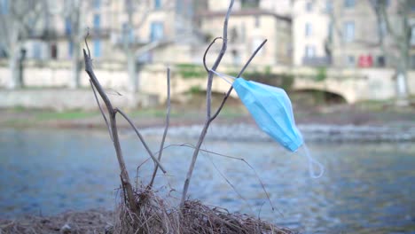 Medical-protective-face-mask-discarded-on-a-branch-next-to-a-river,-blown-by-the-wind,-nature-pollution-during-the-Coronavirus-Covid-19-pandemic