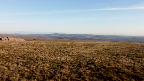 Aerial-Drone-Shot-Moving-Over-Grass-Mountain-With-Wind-Farm-and-Fields-in-the-Distance