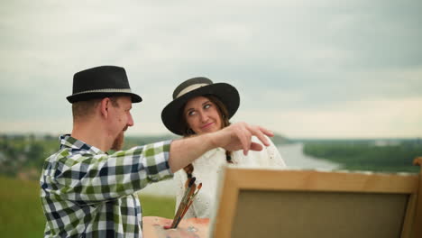 a man in a black hat and plaid shirt stands close to a woman in a white dress in a scenic field. he gestures in a direction, possibly discussing their next steps or an art composition