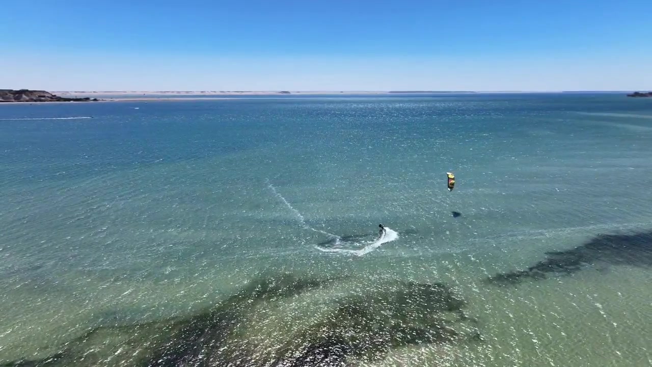 モロッコのダフラ湾の透き通った水面をカイトボーダーが滑走するドローン空撮。ターコイズブルーの浅瀬、海の質感、そして世界クラスのウィンドスポーツが織りなす息をのむような海岸の景色。