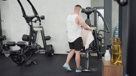 sportsman walks to chair placing sports towel on seat backrest next to table with water bottle and spray preparing workout environment in modern gym with sleek equipment under bright lighting