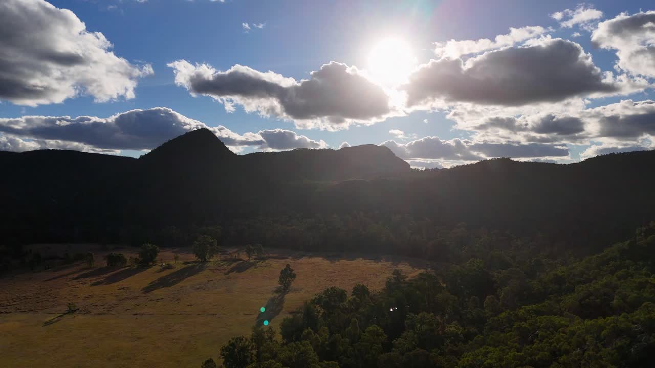 Aerial view of sun setting behind mountain ridge, casting dramatic light over rolling hills and forest in rural New South Wales, Australia