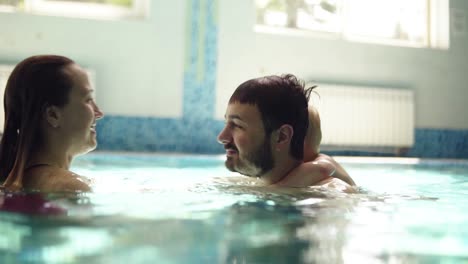 Happy-caucasian-family-of-three-are-hanging-out-together-in-the-swimming-pool.-Happy-wife-is-kissing-her-husband.-Their-little-blonde-son-holds-the-neck-behind-on-the-fathers-back.-Indoors