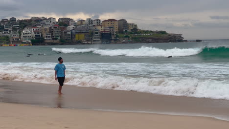 indian man walking barefoot in the bondi beach with surfers suring in the ocean in australia