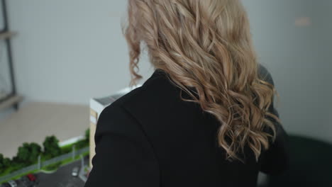 focused businesswoman in black blazer carefully carries architectural building model through office, approaching her desk with wall-mounted board in background