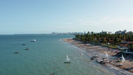 Hochschwenkbare-Weitwinkelaufnahme-Des-Tropischen-Bessa-Strandes-In-Der-Hauptstadt-Joao-Pessoa,-Paraiba,-Brasilien,-Mit-Menschen,-Die-Das-Meer-Genießen,-Kleinen-Fischerbooten-Und-Wolkenkratzern-Im-Hintergrund