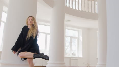 donna bionda concentrata in pullover sciolto a maniche lunghe nere e stivali che eseguono una danza contemporanea in studio