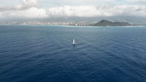 4k drone video captures a lone sailboat drifting peacefully across the deep blue ocean in hawaii. the sailboat glides effortlessly, framed by the rugged green mountains rising in the distance.