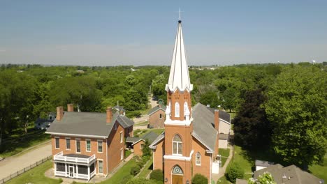 aerial establishing shot of traditional white steeple church behind green trees in summer