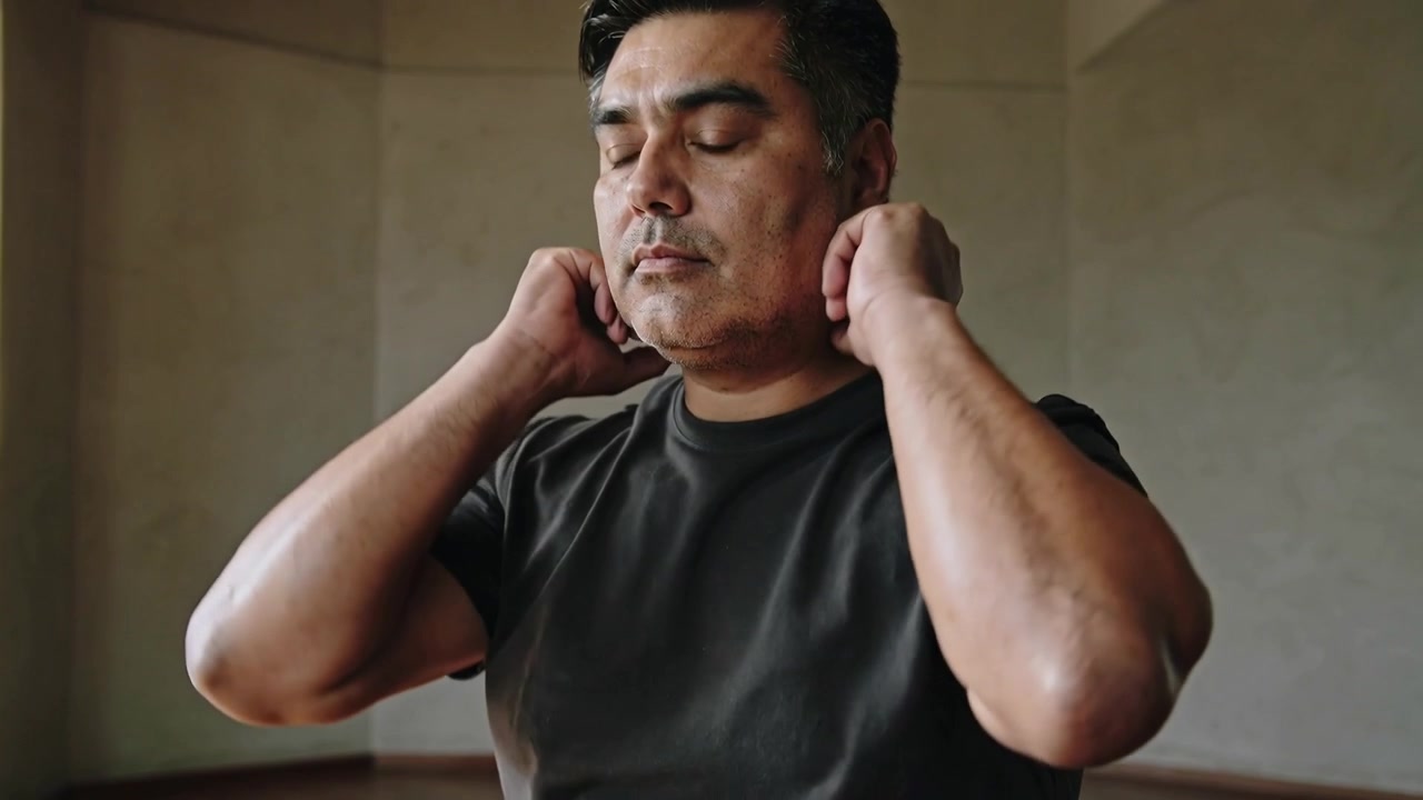 Shifting camera, middle-aged man in dark T-shirt lifting hands and holding neck at home, rolled mat