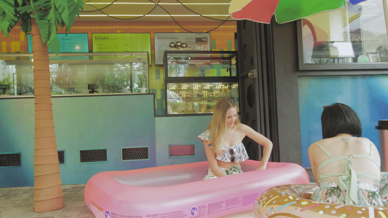 Two young women enjoy ice cream in a relaxed atmosphere next to an inflatable pool in front of a contemporary ice cream parlor.