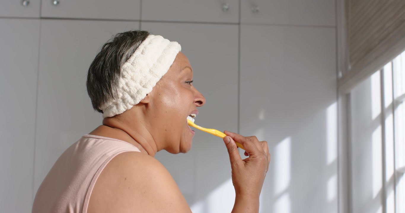 African American woman holding yellow toothbrush near mouth starting brushing teeth in bathroom