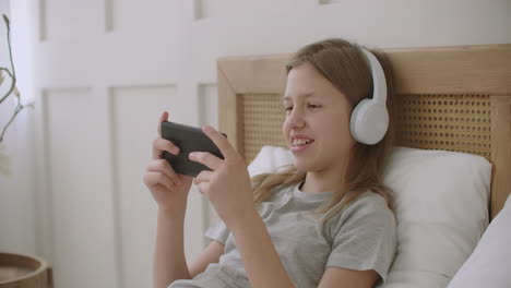 cheerful little girl is lying on bed with smartphone in hands and playing game spend time in internet