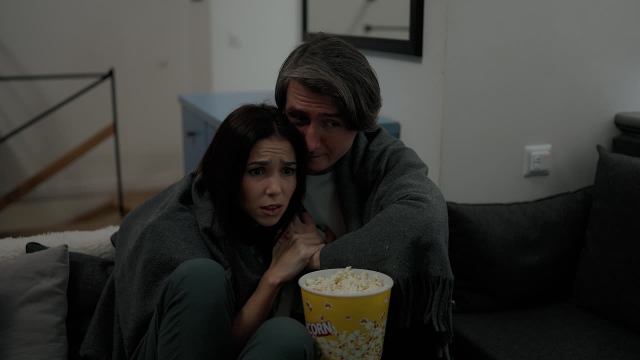 una pareja caucásica ve una película de terror en el sofá y come palomitas de maíz en casa.