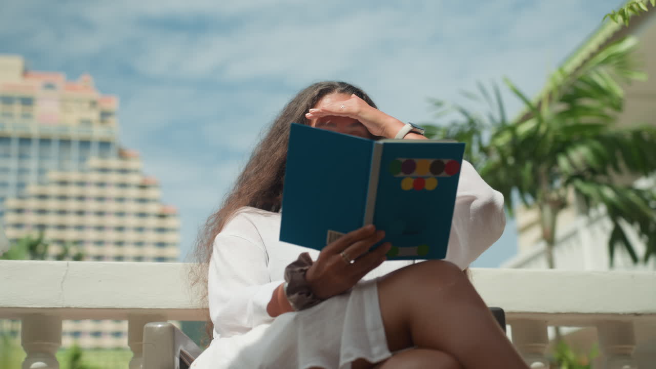 Woman reading outdoors. Woman in white enjoying sunny balcony read. Gentle breeze as woman studies travel guide outside. Peaceful vacation scene with woman reading guide beneath palm trees