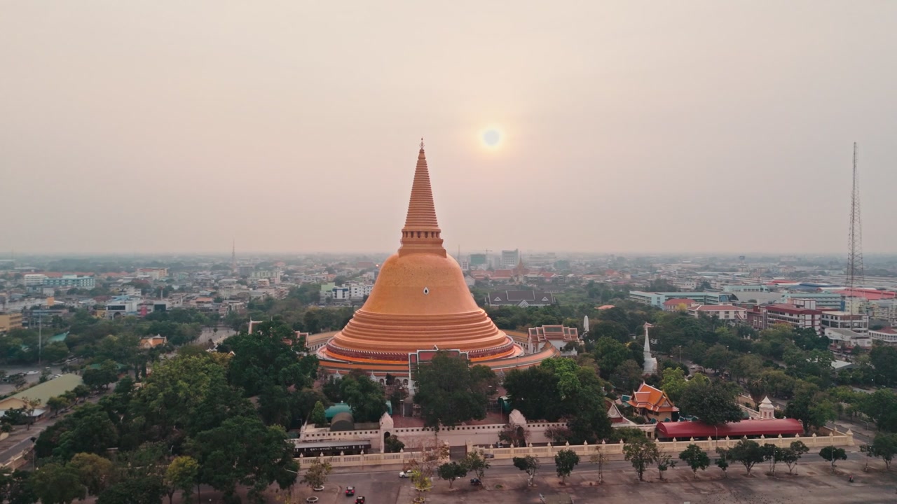 Prospettiva aerea da drone che cattura un enorme stupa dorato che si erge sopra il centro urbano, mentre un maestoso monumento buddista domina lo skyline della città in Thailandia sotto un cielo grigio e velato con un sole splendente.