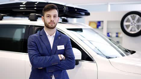 Portrait-of-young-car-salesman-at-car-showroom,-standing-with-crossed-arms