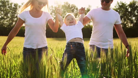 una familia joven con un niño pequeño corriendo en un campo con espigas de trigo balanceando a un niño en sus brazos que ríe y sonríe de felicidad
