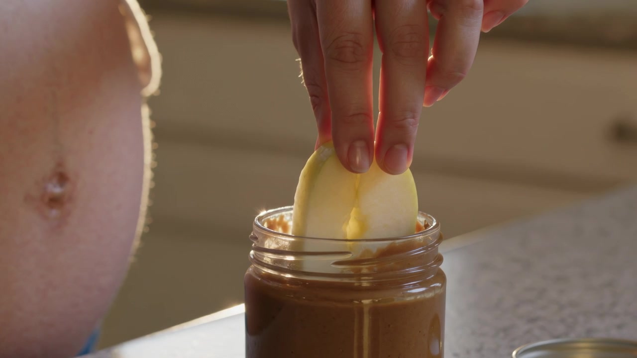 Lowering woman wearing crop top pressing apple slice into spread jar in kitchen, preparing snack