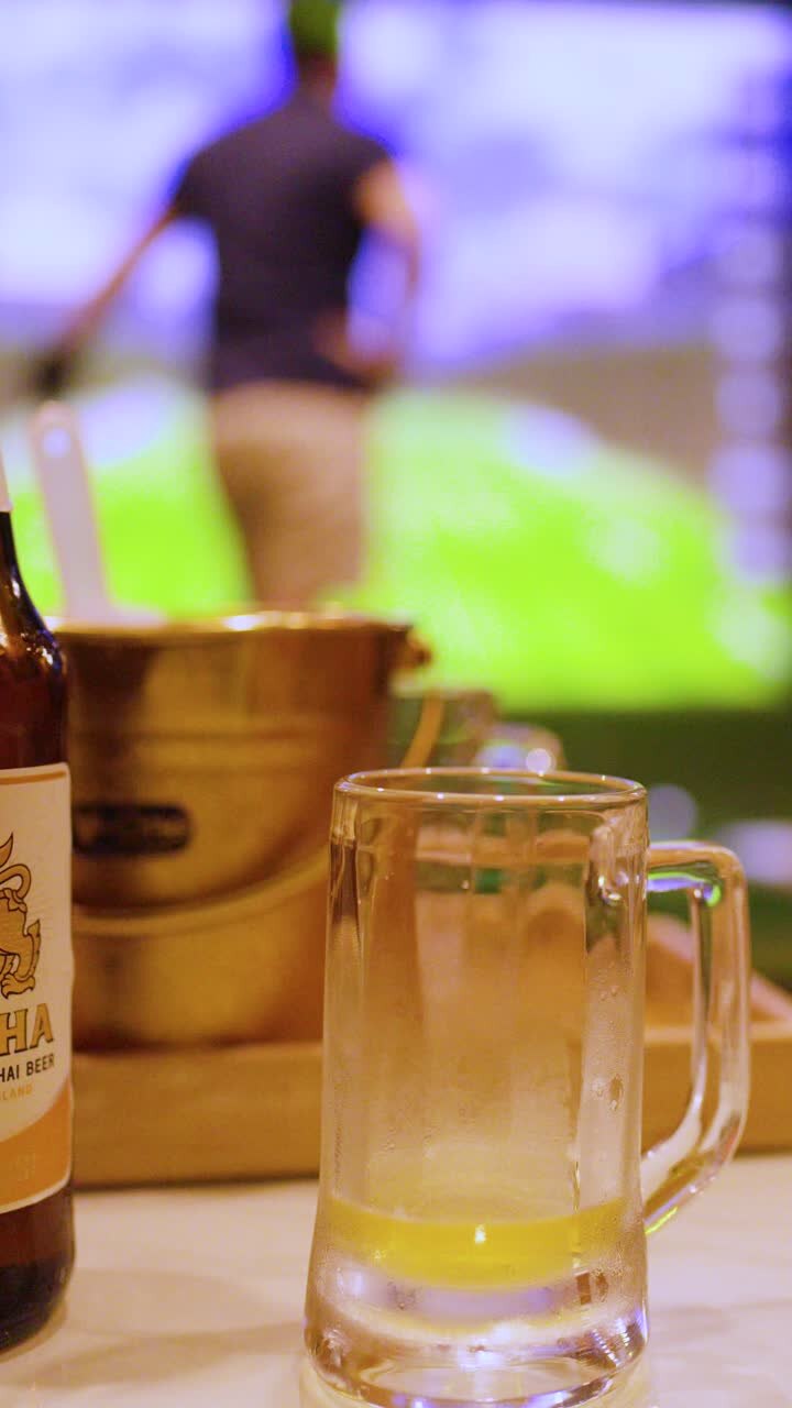 Una bottiglia di birra e un boccale di vetro sono su un tavolo davanti a un simulatore di golf indoor