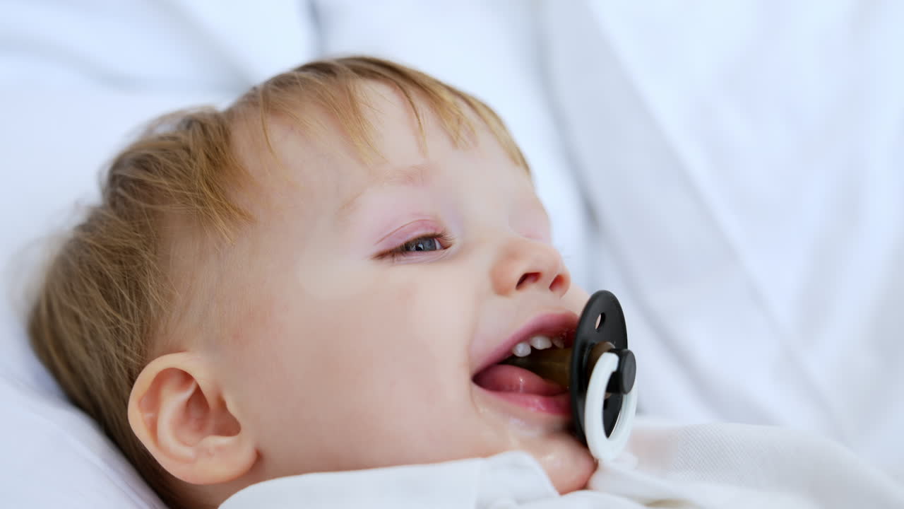 Face of a happy Caucasian blond toddler boy sucking a pacifier. Portrait of a kid falling asleep. Close up.