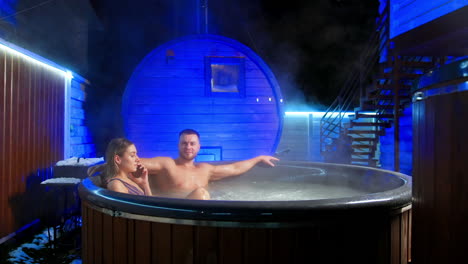 people are relaxing sitting ibn the hot tub outdoors. man sits spread his hands to the sides and woman holds a phone at her ear.