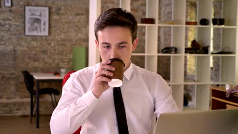 joven empresario está trabajando con una computadora portátil y bebiendo café en la oficina, mirando a la cámara, concepto de negocio, concepto de comunicación