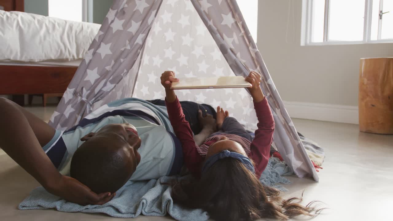 Video di un padre e una figlia afroamericani felici sdraiati in tenda e che usano un tablet insieme