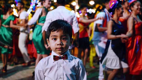 young boy wearing a bow tie stands still in the foreground, contrasting with a vibrant celebration of dancing people in colorful costumes behind him