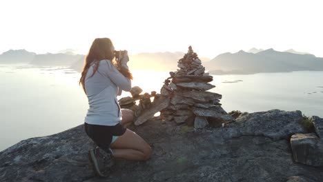 una mujer joven con el pelo largo está sentada y toma una foto con su cámara de un pequeño montón de rocas una encima de la otra