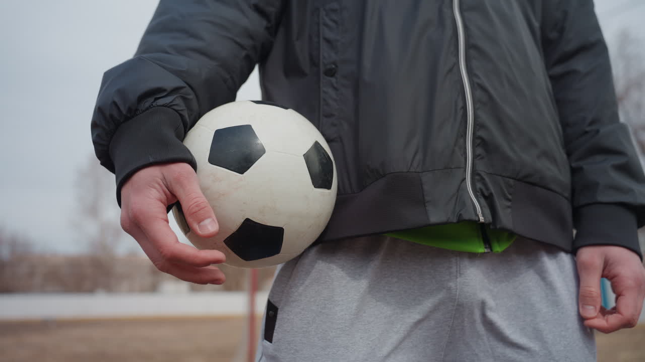 Снятый с полусреднего расстояния спортсмен держит футбольный мяч на бедре. Белый уличный спортсмен в повседневной одежде готовится к обычному матчу на потрепанном местном поле. Едва заметное напряжение в пальцах и позе говорит о готовности.