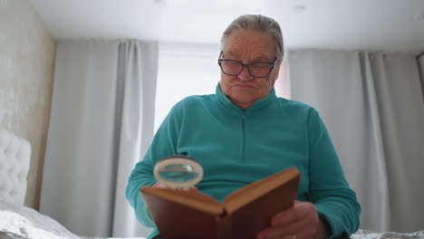 elderly woman in teal shirt sitting on bed holding book, deeply focused on reading with magnifying glass, looking puzzled or confused. bright, calm indoor setting with soft light