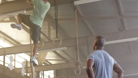 diverse male coach watching man training at gym climbing rope, in slow motion