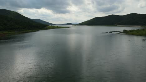 aerial footage towards the horizon revealing mountains, the dam, a bird passing in front of the camera, the water that reflects the sun setting, muak klek, saraburi, thailand