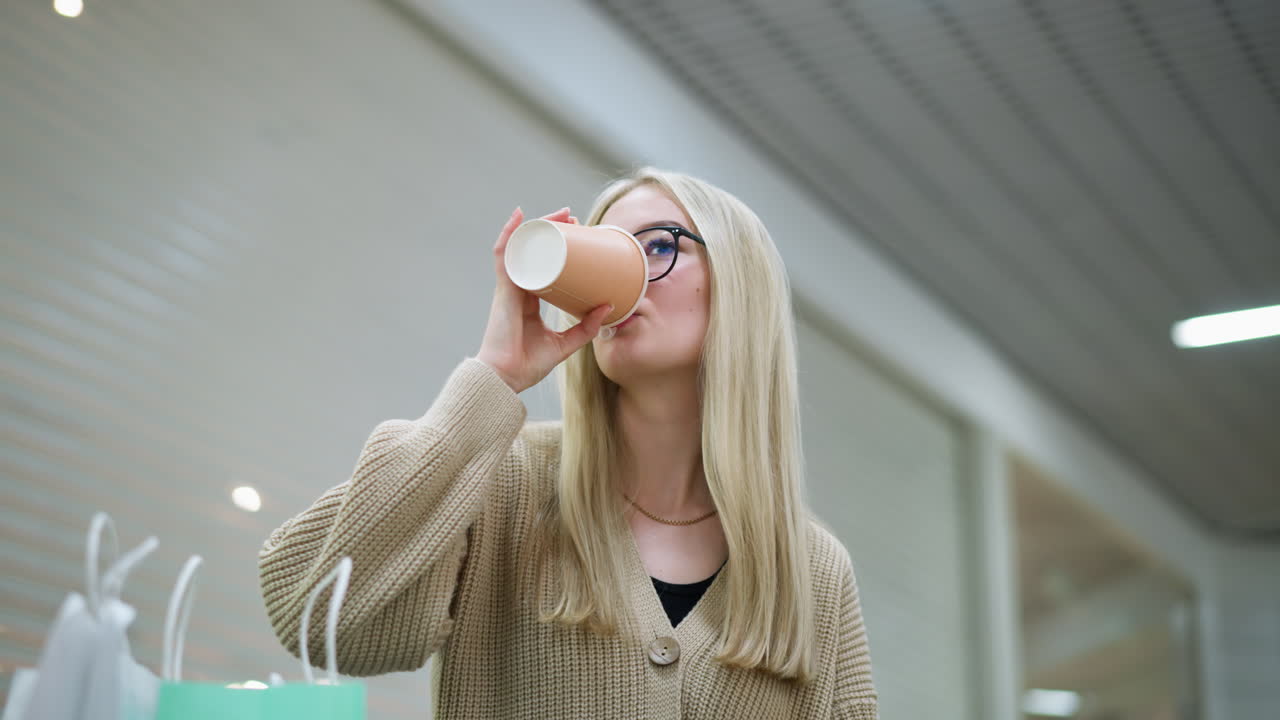 dama con suéter de punto y gafas beba un sorbo mientras dos bolsas de compras se sientan cerca en un centro comercial bien iluminado, el fondo presenta luces de techo brillantes y un entorno comercial moderno