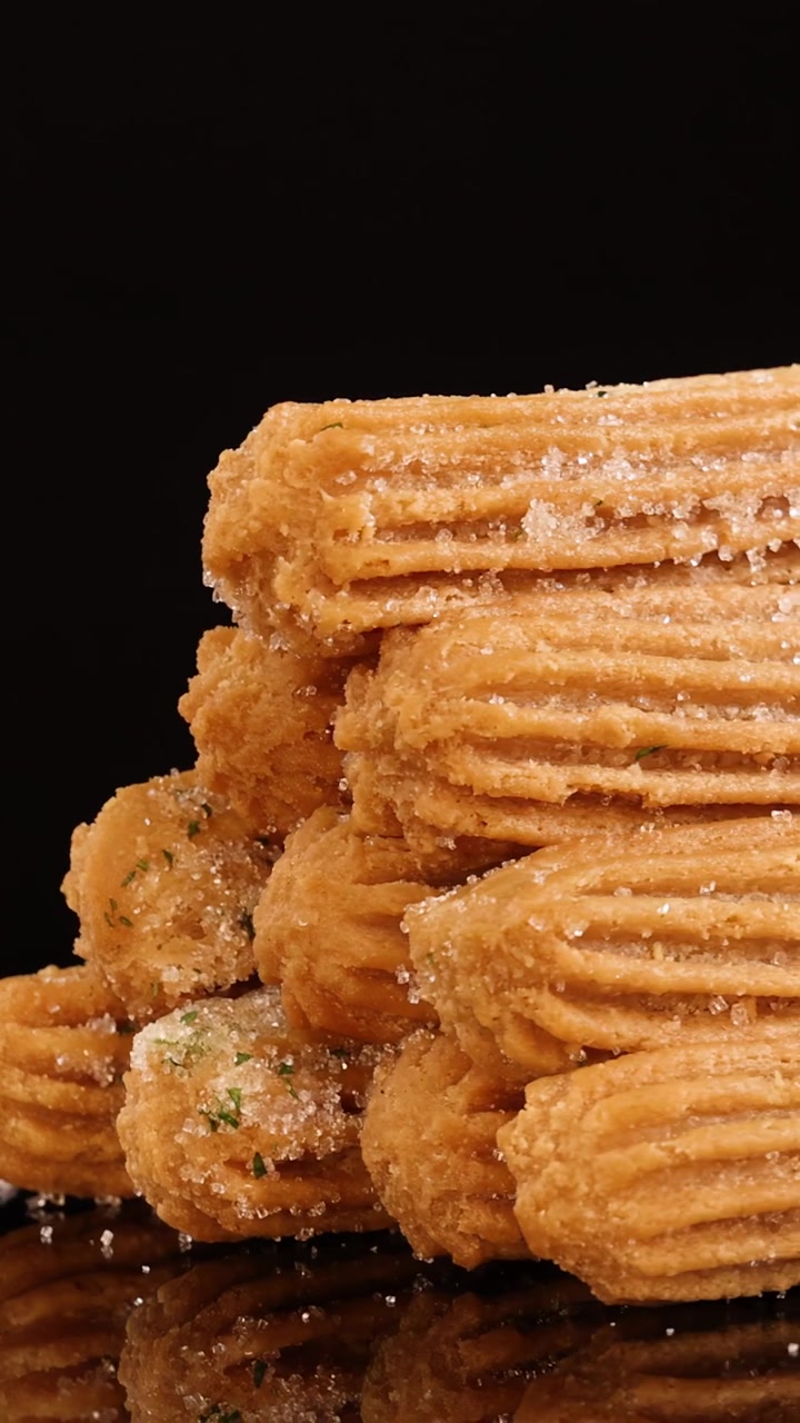 Golden churros coated with sugar rotate smoothly on a reflective black surface, illuminated by soft studio lighting for a crisp, appetizing appearance