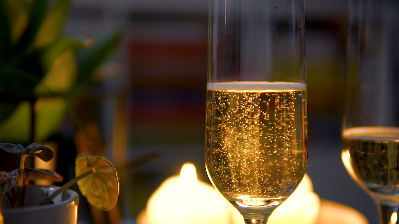 Primo piano di champagne dorato in un calice da flute con bollicine che salgono, sullo sfondo di luci di candele sfocate sul tavolo.