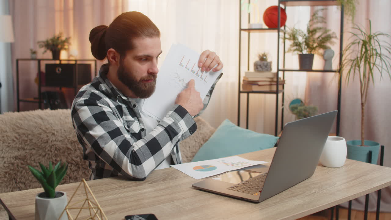Young man making laptop video chat at home table showing paperwork through webcam positive feedback