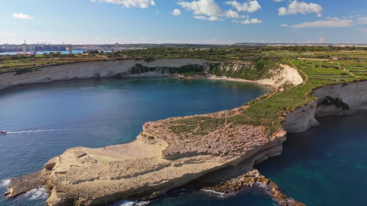 Vista aerea di una tranquilla baia maltese con imponenti scogliere calcaree e acque turchesi del Mediterraneo, con Marsaxlokk visibile sullo sfondo sotto un cielo blu brillante
