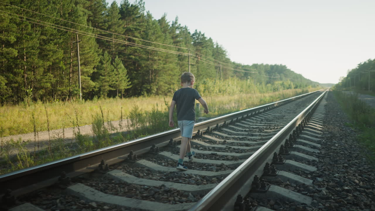 rear view of young boy in dark tee and light shorts walking carefully on rail track surrounded by gravel and wild grass with forest background under clear sky and warm sunlight casting soft glow