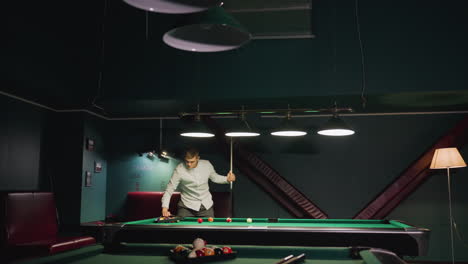 player in beige shirt leans over green pool table, focusing on shot. dimly lit room with dark walls creates relaxed atmosphere for game of billiards or pool