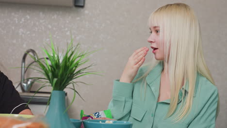 young lady licking fingers smiling while talking to friend in kitchen setting, natural moment of enjoyment and conversation, snacks and green plant on table, casual home atmosphere