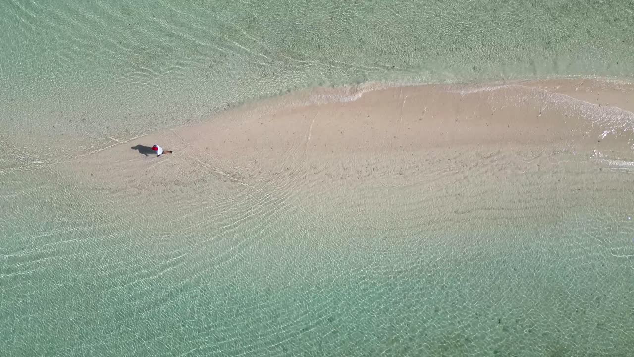 One person wading in crystal clear lagoon towards beached boat as water lapping on thin sandbar, Drone overhead follow shot