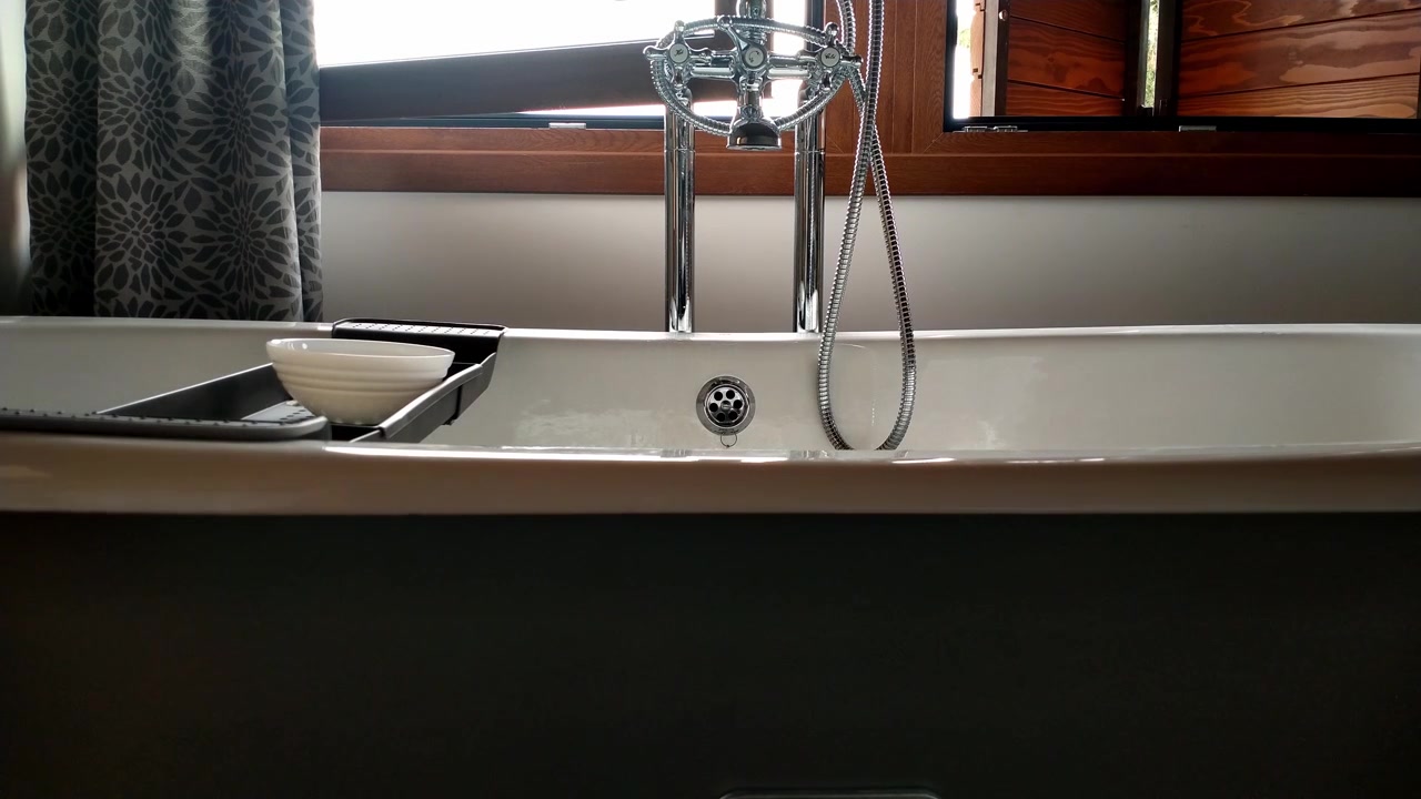 Hand turns on water tap in modern bathtub by window with natural light and curtain