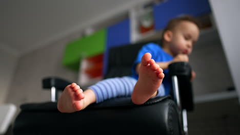 toddle sitting in the chair his bare feet close to camera. little child leans in the chair looking down. blurred backdrop. low angle view.